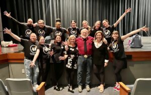 Group on stage wears black "ARTSPARKS DANCE. MUSIC. JOY" shirts surrounding a man in red.