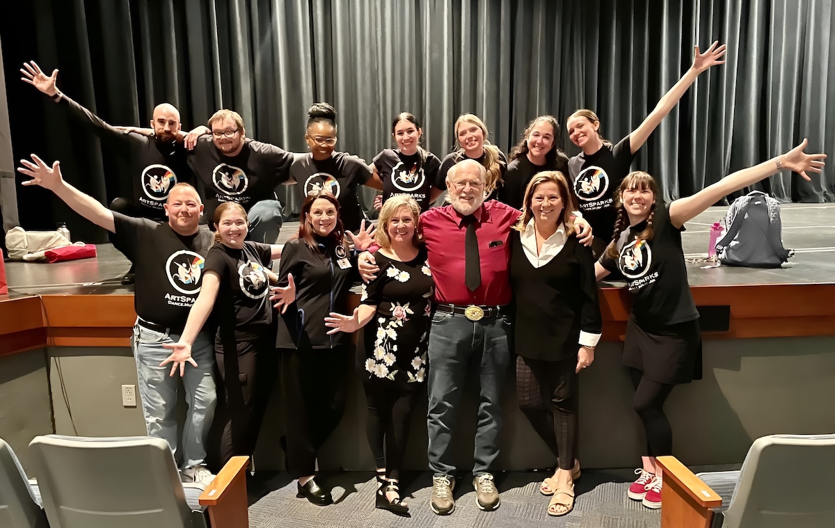 Group on stage wears black "ARTSPARKS DANCE. MUSIC. JOY" shirts surrounding a man in red.
